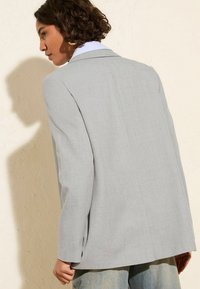 Světle šedý blazer s texturovanou tkaninou, designem na jednobutton a klopovými límci. Má minimální prošití a mírně převislé rukávy.