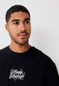 Camiseta de algodón negra con cuello redondo que presenta "Felices Fiestas" en letra blanca, enmarcado por un bordado de luces navideñas coloridas.