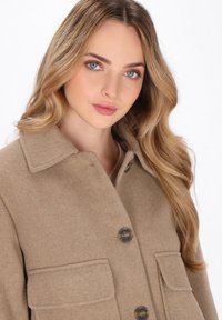Cappotto in misto lana color beige con colletto, chiusura frontale con bottoni e due tasche applicate, caratterizzato da una consistenza morbida e dettagli di cucitura sottili.