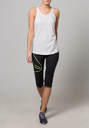 Žena v bílém tílku s pruhy, černých capri legínách se žlutým vzorem a černých běžeckých botách kráčí vpřed na jednobarevném pozadí.