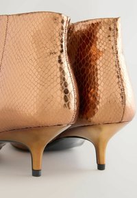 Bottines en cuir couleur cuivre avec une texture en effet serpent, dotées d'un talon effilé et d'une finition métallique lisse, accentuées par une surface brillante.