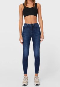 Jeans skinny azul oscuro con una textura de denim suave, que presentan una cintura alta y detalles de franjas laterales, combinados con una camiseta corta negra.