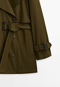 Oliwkowy trencz z podwójnym zapięciem, paskiem w talii, dużymi guzikami oraz bocznymi kieszeniami. Gładki, trwały materiał.