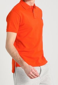 Orange polo-shirt med krage och korta ärmar. Tillverkad av bomull, har sidofickor för ökad rörelsefrihet. Inga synliga logotyper eller mönster.