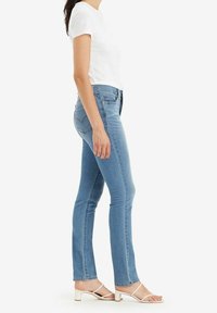 Nainen vaaleansinisissä slim-fit-farkuissa, valkoisessa lyhythihaisessa paidassa ja valkoisissa avokkaissa seisoo sivuttain valkoista taustaa vasten.