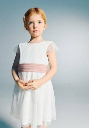 Niña joven con cabello rojo, vestida con un vestido blanco con una faja rosa, de pie con las manos ligeramente entrelazadas frente a un fondo gris neutro.