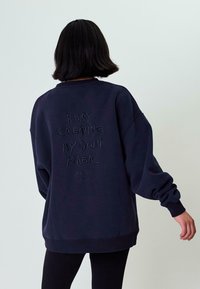 Felpa navy con vestibilità oversize. Presenta un ricamo sul retro con la scritta "STO CREANDO LA MIA MAGIA" in un carattere testurizzato e giocoso.
