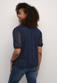 Blusa corta a maniche corte di colore blu navy con un intricata lavorazione in pizzo, caratterizzata da un motivo a diamante e maniche leggermente a palloncino. Texture liscia.