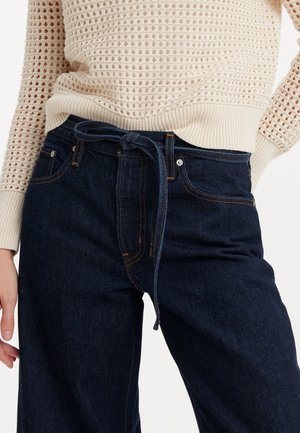 Person iført mørkeblå jeans med et bundet denimbælte og en cremefarvet åben strikket sweater, hånd hvilende nær låret.