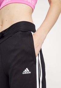 Schwarze Sporthose mit weißen Streifen, ausgestattet mit einem Kordelzug und Seitentaschen. Adidas-Logo deutlich auf dem linken Bein angebracht.