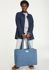 Sac fourre-tout en tissu bleu avec deux poignées, de forme rectangulaire, en tissu texturé, étiquette à l'avant, associé à une veste en jean et un vêtement de détente gris.