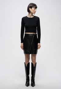 Svart croppad långärmad topp kombinerad med en svart läderkjol med diagonal dragkedja och en slät yta, kompletterad med knähöga svarta stövlar.