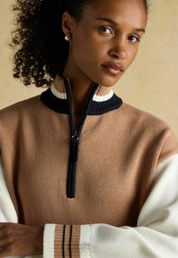 Kortsukelatega naine kannab pruun-valget poolsiidi, millel on mustvalge triibuline krae ja sobivad triibulised mansetid, beežil taustal.