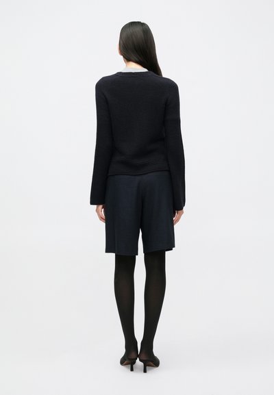 Sötét kötött pulóver hosszú ujjakkal, sötét szabott rövidnadrág felett viselve, átlátszatlan fekete harisnyával és fekete magassarkú cipővel párosítva.