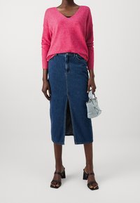 Różowofuksjowy sweter z dekoltem w serek, dopasowana spódnica dżinsowa z rozcięciem z przodu, czarne sandały na słupku oraz mała jasnoniebieska torebka z akcentami.