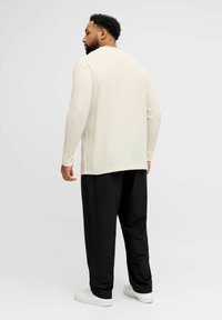 Lys beige, langermet skjorte med teksturert stoff, kombinert med vide svarte bukser og hvite joggesko, sett bakfra.