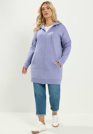 Frau trägt einen langen lavendelfarbenen Kapuzenpullover mit Reißverschluss, blaue hochgekrempelte Jeans und weiße Sneaker und steht vor einem schlichten hellen Hintergrund.