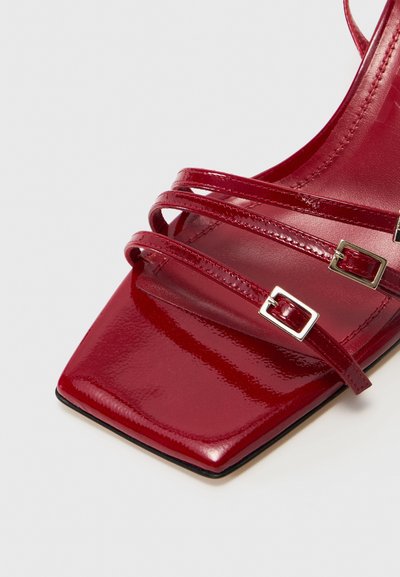 Czerwone lakierowane sandały na wysokim obcasie z kwadratowym noskiem, wieloma skrzyżowanymi paskami i srebrnymi akcentami na klamrach pasków.