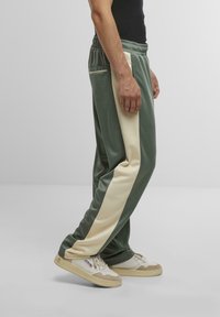 Grüne und cremefarbene Sweatpants mit glatter Textur, ausgestattet mit Seitentaschen und einem entspannten Schnitt, kombiniert mit hellen Sneakern.