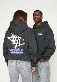 Zwart hooded sweatshirt met witte en paarse grafische ontwerpen. Heeft een ritssluiting aan de voorkant, een losse pasvorm en ribgebreide boorden. Draagt met grijze jeans.