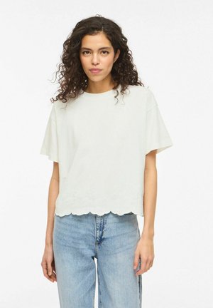 Femme aux cheveux bouclés portant un haut blanc à manches courtes avec bord ondulé et un jean taille haute bleu clair, debout devant un fond blanc.