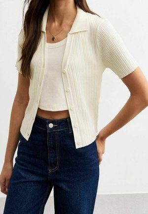 Woman wearing a white ribbed short-sleeve buttoned cardigan over a white cropped tank top and high-waisted dark blue jeans.