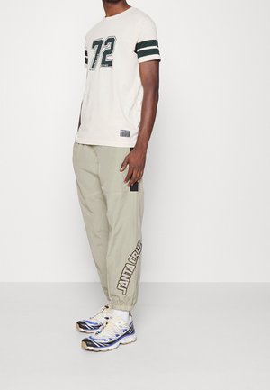 Beige t-shirt met groene strepen en nummer 72. Lichtgrijze cargo broek met een zwart patch en "SANTA CRUZ" in bruin gedrukt. Sportieve sneakers.