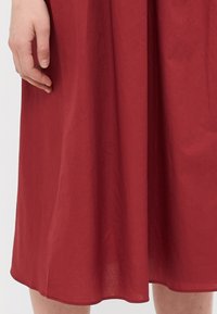 Jupe rouge mi-longue en tissu léger ; texture lisse avec un léger brillant ; poche sur la couture latérale visible ; l'ourlet a une doux tombé.