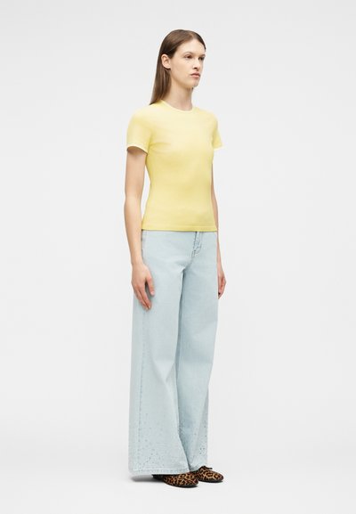 Γυναίκα στέκεται κοιτώντας προς τα δεξιά, φορώντας ένα στενό κίτρινο κοντομάνικο πουκάμισο, ανοιχτόχρωμα τζιν με φαρδιά πόδια και λεοπάρ επίπεδα παπούτσια σε λευκό φόντο.