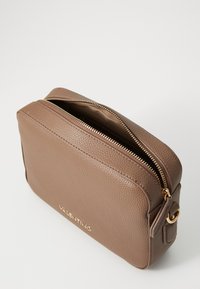 Bruine leren cosmetica tas met een gestructureerd oppervlak, gouden rits en logo-detail. Heeft een rechthoekige vorm met afgeronde hoeken.