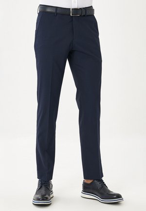 Uomo con pantaloni eleganti aderenti blu navy, cintura nera e scarpe brogue nere su sfondo bianco.