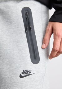 Pantaloni de trening gri cu o textură netedă, având o buzunar cu fermoar de un gri închis proeminent și logo-ul negru Nike în colțul din stânga jos.