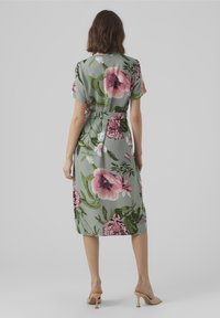 Donna in piedi che indossa un vestito midi verde con una grande stampa floreale rosa, maniche corte, vita cintata e mules beige con tacco, vista da dietro.