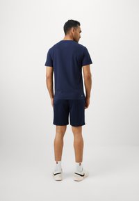 Άνδρας με το πρόσωπο στραμμένο μακριά, φορώντας μπλέ μπλουζάκι και σορτς, λευκά αθλητικά παπούτσια Nike και λευκές κάλτσες με κόκκινο και μαύρο λογότυπο.