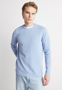 Maglietta azzurra chiara a maniche lunghe con texture a coste e scollo rotondo, con un piccolo logo sul petto. Indossata con jeans chiari.
