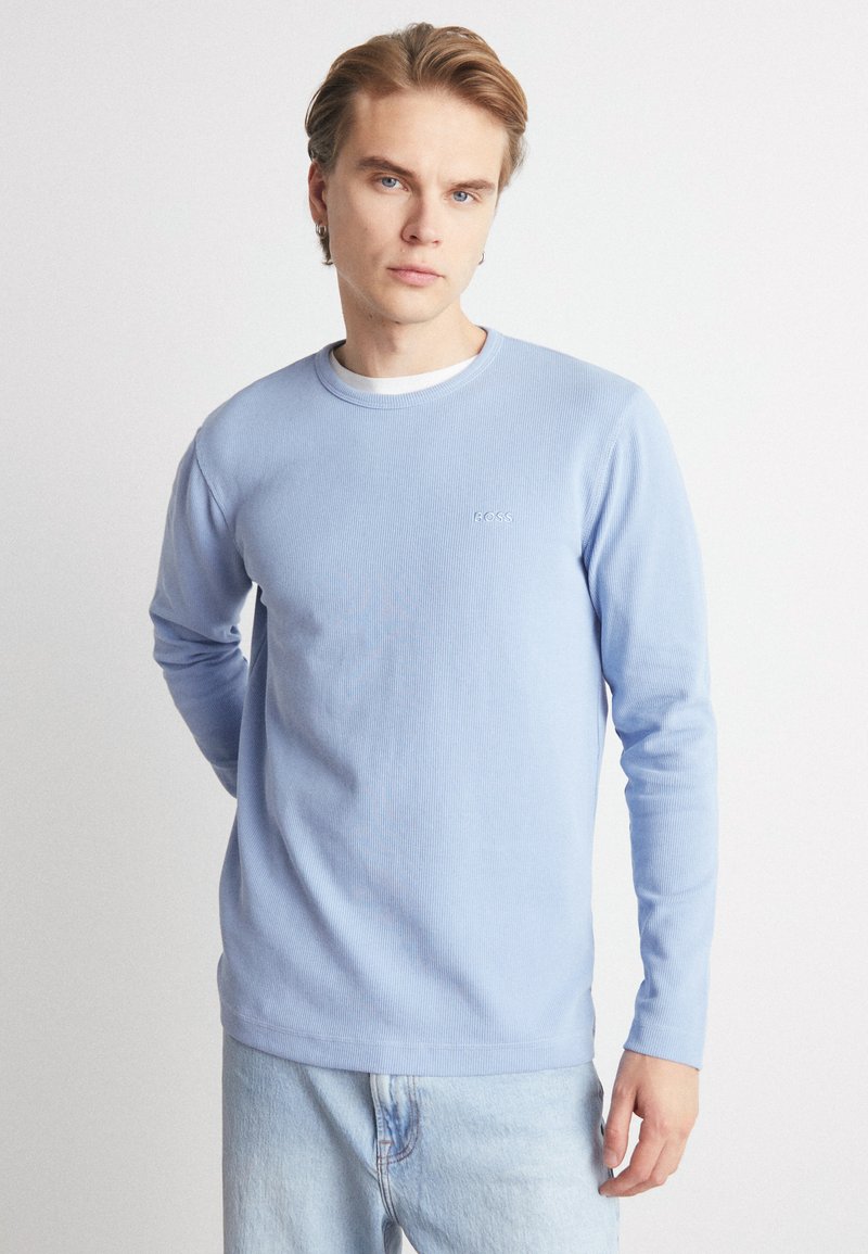 Maglietta azzurra chiara a maniche lunghe con texture a coste e scollo rotondo, con un piccolo logo sul petto. Indossata con jeans chiari.