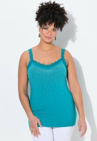 Top de tirantes color teal con encaje y un brillante patrón de piedras de strass en el frente, hecho de una tela suave, combinado con pantalones blancos.
