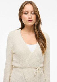 Krēmkrāsas wrap-stila kardigans, izgatavots no mīksta, pūkaina auduma. Tam ir V-veida izgriezums, garas piedurknes un josta pie vidukļa.