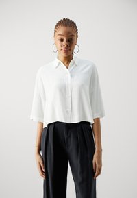 Camicia bianca, corta con bottoni e maniche corte, abbinata a pantaloni neri a vita alta con pieghe. Tessuto liscio, design minimale.