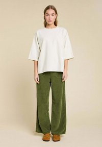 Un haut crémeux oversize avec des manches larges, associé à un pantalon large en velours côtelé vert. Des chaussures marron à enfiler complètent le look.