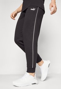 Calças de desporto pretas com riscas laterais brancas, feitas de um material suave. Combinadas com ténis brancos com um logótipo discreto.