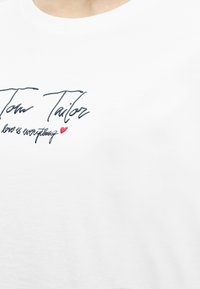 Άσπρο βαμβακερό t-shirt με μαύρο σχέδιο υπογραφής και κόκκινη καρδιά, με στρογγυλή λαιμόκοψη και κοντά μανίκια.