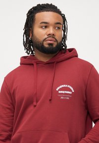 Hombre con barba y dreadlocks vistiendo una sudadera roja con texto blanco en el pecho, mirando de lado frente a un fondo liso.