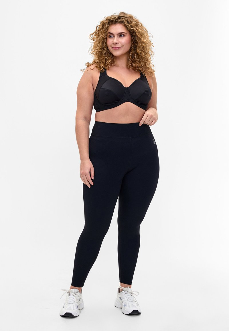 Sort sports bh og højtaljede sorte leggings, der har en glat tekstur og et enkelt design. Modellen har hvide træningssko på.