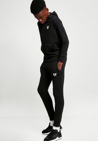 Sudadera negra con capucha y pantalones ajustados de mezcla de algodón. La sudadera cuenta con un bolsillo frontal y un logo; los pantalones tienen bolsillos laterales y un logo a lo largo de la pierna.