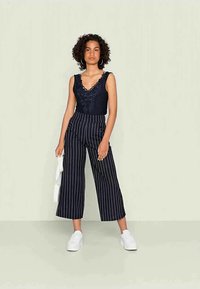 Mulher com cabelo encaracolado vestindo uma blusa preta sem mangas com bordados florais, calças largas listradas em preto e branco, tênis brancos e segurando uma bolsa branca.