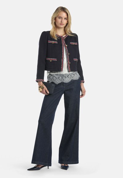 Kvinde iført marineblå cropped jakke med rød og hvid kant, hvid blonder top, vidde bukser i mørk denim, sorte hæle og holder en sort clutch.