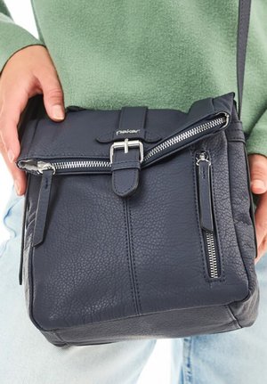 Person hält eine schwarze Leder-Umhängetasche mit silbernen Reißverschlüssen und Schnalle, trägt einen grünen Pullover und hellblaue Jeans.