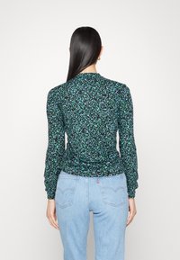 Långärmad blus med ett svart och grönt blommönster, med en rynkad baksida och rundad halsringning. Kombinerad med ljusblå jeans.