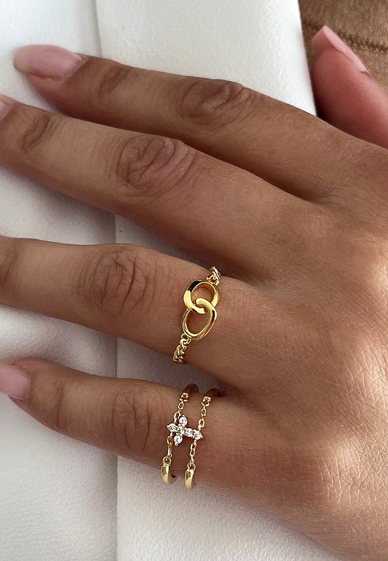 Gouden ringen op een hand; de ene heeft een interlocking lusontwerp, de andere heeft een klein kruis met heldere accenten. Zachte textuur, delicate kettingen.